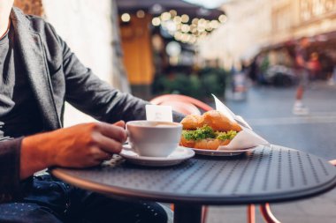 Bir fincan kahveyi erkek ellerinde kruvasanla kapat. Gün batımında açık kafede sıcak kapuçino içen adam terasta masada oturuyor..