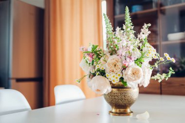Pirinç vazoda yapılmış taze çiçek aranjmanı mutfak dekorasyonu masasının üzerine konmuş. Beyaz bej güllerden yapılmış buket. Dolabın yanında masa takımlarıyla bırakılmış dijitalis çiçeği.