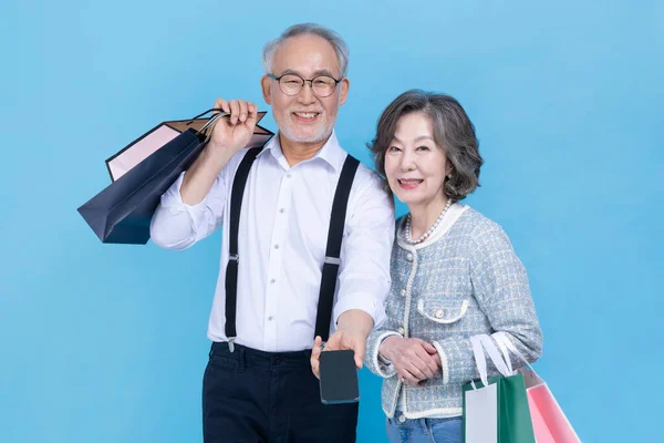 在工作室背景下手持购物袋和智能手机的活跃的资深韩国老年夫妇 — 图库照片