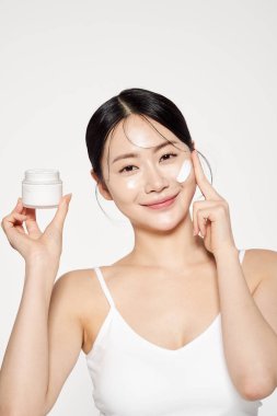 an Asian woman who holds a cream container and applies cosmetics to her face clipart