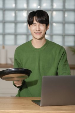 A man in his 20s holding a frying pan and using a laptop clipart