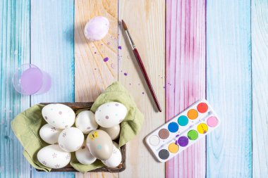 Painted Easter eggs in a basket and elements to paint, on a colorful wooden background.