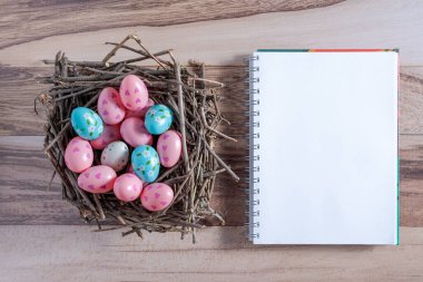 Top view of some Easter eggs painted on a nest of twigs and a blank ciuderno on a wooden background.