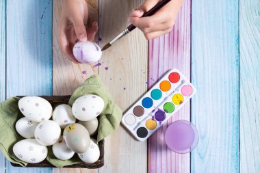 Close-up of a child's hands painting Easter eggs on a colorful wooden background.