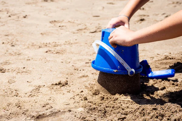 一个在沙滩上玩铲子和蓝色塑料桶的孩子的手 — 图库照片