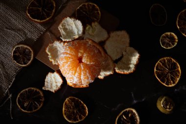 Kurutulmuş turunçgil dilimleriyle çevrili yarım soyulmuş mandalina manzarası. Işıklar ve gölgeler koyu yiyecek tarzı.