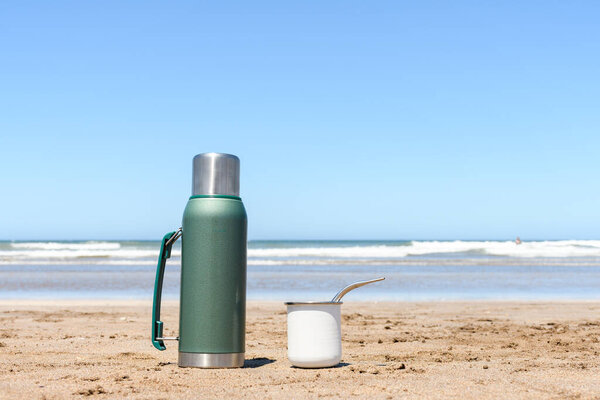Mate and thermos in the sand of the beach in front of the sea, with copy space.