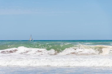 Sahilde dalgalar kopuyor ve arka planda bir yelkenli..