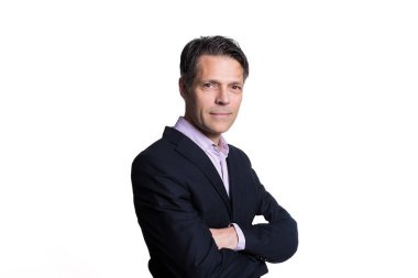 Beyaz arka planda kameraya gülümseyen orta yaşlı bir adam, resmi kıyafetler giyiyor..