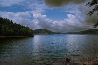 Vosges dağlarında bulutlu bir gökyüzünün altında Pierre Perce Gölü (Pierced Stone Gölü). Göl neredeyse tamamen köknar ormanlarıyla çevrili, vahşi bir manzara yaratıyor..