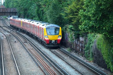 Londra, İngiltere - 30 Mayıs 2019: Çağdaş kırmızı ve sarı yolcu treni yemyeşil bir alanda seyahat ediyor. Tren, Güney Batı Demiryolu Servisi 'nin bir parçası. Etrafı bitki örtüsü ve grafiti kaplı duvarlarla çevrili raylardan geçiyor.