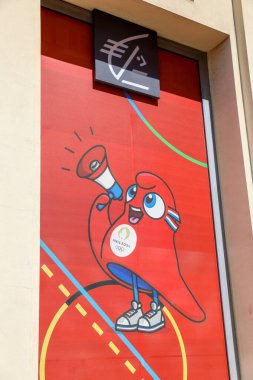 Nice, Fransa - 12 Haziran 2024: Paris 2024 Olimpiyatları 'nın maskotunun yer aldığı renkli bir poster, belirgin bir şekilde bir binada sergilenmektedir. Canlı kırmızı arka plan ve megafonu elinde tutan hevesli maskot yaklaşan megafonun reklamını yapıyor.