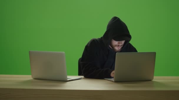 認識できない男のハッカーは ウッドテーブルの上に2台のラップトップコンピュータのキーボードを入力してフードとキャップ付きのスウェットシャツを身に着けています — ストック動画