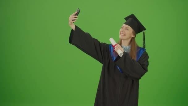 緑の画面 クロマキー 卒業証書と黒ガウンのポーズで笑顔の女性卒業生はセルフィーを作っています 女性学生が修士号を手に男性と帽子をかぶって 大学研究科 — ストック動画