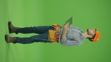 Dikey Görüntü. Yeşil Ekran. Yan Görünüm. Genç Sakallı İnşaat Mühendisi Kareli Gömlek, Güvenlik Miğferi ve Deri Alet Kemeri Giyiyor Etrafa Bakınıyor ve Tablet 'teki Kayıtları Düzeltiyor