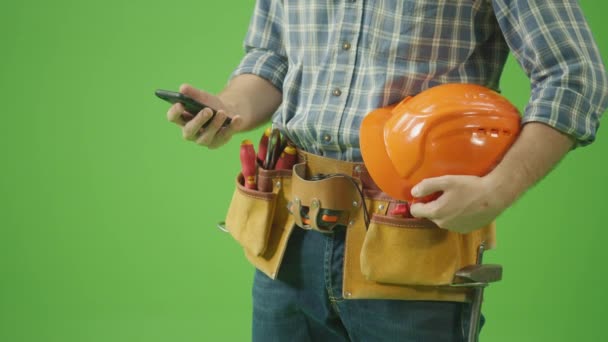 閉鎖中だ 緑の画面 レザーツールベルトと作業手袋とチェックシャツの男は ハード帽子を保持し 他の手で彼の腕とスクロール電話の下にあります 現代の労働者と近代的な建設コンセプト — ストック動画