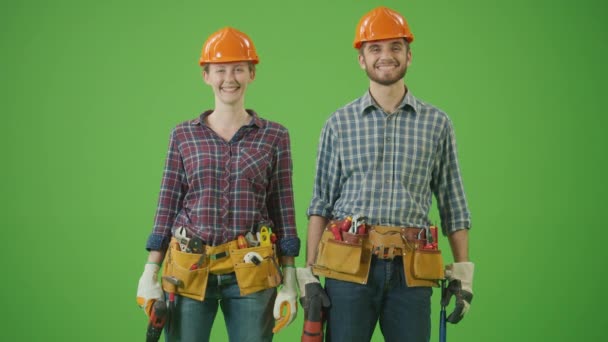 绿色屏风 年轻男女土木工程师穿着格子呢衬衫 工作服 工具带 起跳和钻钻 背对背朝前看相机微笑 — 图库视频影像