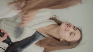 Dikey Video.Gülümseyen Kadın 'ın Arka Görünümü Saçını Aynanın Önünde Kurutuyor. Bornozlu mutlu bir genç kadın saçını yıkadıktan sonra saç kurutma makinesine üflüyor, onu şekillendiriyor. Saç ve Güzellik Konsepti.