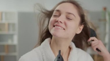 Gülümseyen bir kadının portresi saçlarını aynanın önünde kurutur. Bornozlu mutlu bir genç kadın saçını yıkadıktan sonra saç kurutma makinesine üflüyor, onu şekillendiriyor. Sağlıklı saç konsepti..