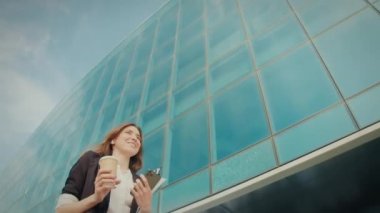 Cep telefonuyla konuşan mutlu iş kadını. İş yerinin önünde yürürken kulaklıkla elinde kahve fincanı tutuyor. İnsanlar ve Uygulamalar. Teknoloji ve İş