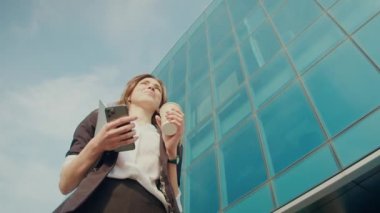 Cep telefonuyla konuşan mutlu iş kadını. İş yerinin önünde yürürken kulaklıkla elinde kahve fincanı tutuyor. İnsanlar ve Uygulamalar. Teknoloji ve İş