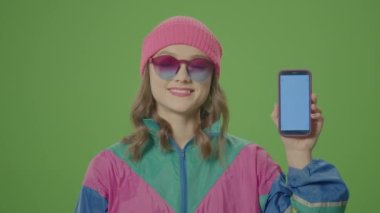 Yeşil Ekran. 80-90 'ların son derece mutlu, pembe şapkalı ve güneş gözlüklü kızı. Mavi Telefondaki parmağıyla işaret ediyor. Dijital Jenerasyon, Genç Nesil ve Teknoloji Konsepti