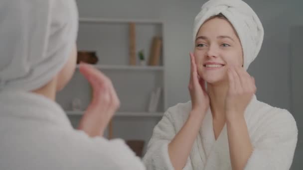 笑顔の女性のバックビューは ミラーを見て 彼女の顔にクリームを適用します ホームで彼女の肌についての彼女の頭のケアに白いタオルを持つ幸せな若い女性 現代の化粧品技術 — ストック動画