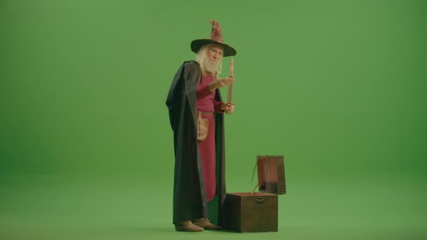绿色屏风 一个穿着中世纪衣服和魔法帽的老魔法师从魔法的胸膛中拿出了魔法之剑 魔法物品 魔法盒 镜子和其他艺术品 — 图库视频影像
