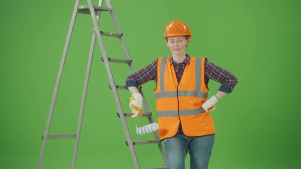 绿色屏风 年轻快乐自信的女建筑工人 穿着格子衬衫 安全夹克 助听器 靠在梯子上 提起油漆滚筒 做衬垫和微笑 — 图库视频影像