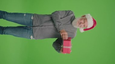 Dikey Görünüm. Yeşil Ekran. Noel Baba Şapkalı Yaşlı Bir Adamın portresi Kırmızı Hediye Kutusuyla. Yaşlı bir adam hediyeyi inceler ve başparmağını kaldırır. Mutlu Noeller Sevkiyat Teslimatı.
