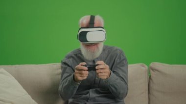 Yeşil Ekran. VR gözlüklü yaşlı bir adam, oyun çubuğuyla bilgisayar oyunu oynuyor, koltukta oturuyor. Yaşlı bir adam Playstation oynuyor ve kazanıyor. Teknoloji Nostaljisi ve Nesil Bölümü.