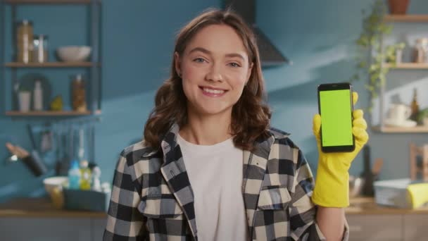 厨房里一个身穿黄色橡胶护目镜的微笑的年轻女子的画像 一个女家庭妇女拿着一个带着绿色屏风的智能手机 在他面前展示 智能清洁技术 — 图库视频影像