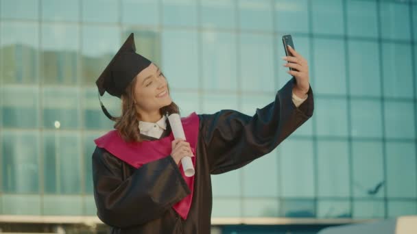 卒業証書と黒ガウンのポーズで笑顔の女性卒業生はセルフィーを作るか ビデオコールを持っています 女性学生が修士号を手に男性と帽子をかぶって 大学研究科 — ストック動画