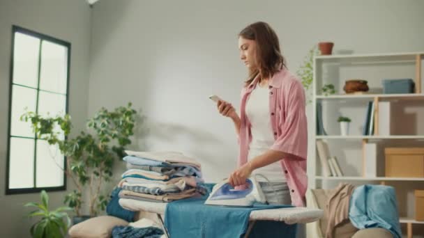 Втомлена Домогосподарка Прасує Одяг Вдома Жінка Використовує Свій Телефон Тому — стокове відео