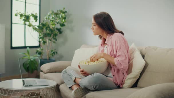 一位年轻女子吃爆米花 专心致志地在笔记本电脑上看电影 一个严肃的女人坐在沙发上看一部非常有趣的电影 电影类型和情感反应 — 图库视频影像