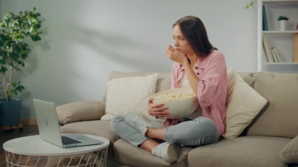 一位年轻女子吃爆米花 专心致志地在笔记本电脑上看电影 一个严肃的女人坐在沙发上看一部非常有趣的电影 电影类型和情感反应 — 图库视频影像