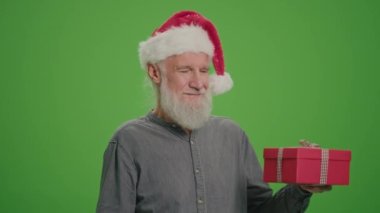Yeşil Ekran. Noel Baba Şapkalı Yaşlı Bir Adamın portresi Kırmızı Hediye Kutusuyla. Yaşlı bir adam hediyeyi inceler ve başparmağını kaldırır. Mutlu Noeller Sevkiyat Teslimatı.