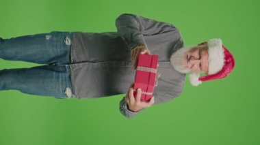 Dikey Görünüm. Yeşil Ekran. Noel Baba Şapkalı Yaşlı Bir Adamın portresi Kırmızı Hediye Kutusuyla. Bir Yaşlı Adam Sınavlar ve Bir Hediye Verir. Mutlu Noeller Sevkiyat Teslimatı.
