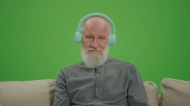 Yeşil Ekran. Gri Sakallı Mutlu Bir Yaşlı Adam Kulaklıkla Müzik Dinliyor, Koltukta Oturuyor. Yaşlılar için Gelişen Teknolojiler.