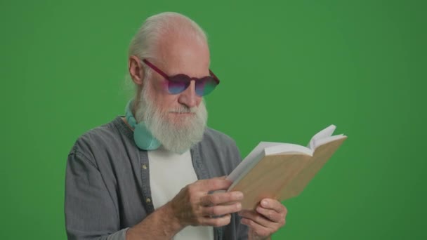 绿色屏风 一个长着灰胡子的聪明老人的画像 有趣的眼镜和耳机 一个老年人翻阅着一本书的书页 康乐活动 康乐活动概念 — 图库视频影像