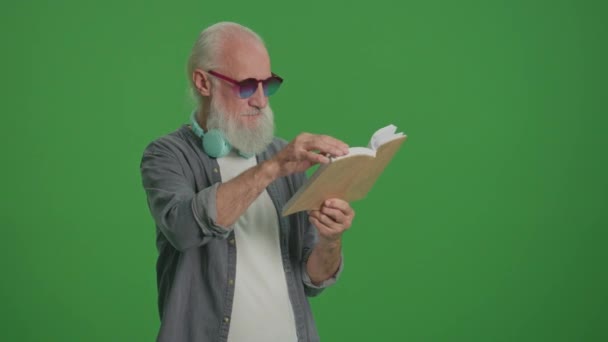 绿色屏风 一个长着灰胡子的聪明老人的画像 有趣的眼镜和耳机 一个老年人翻阅着一本书的书页 康乐活动 康乐活动概念 — 图库视频影像