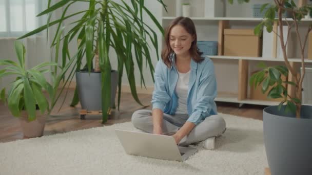 笑顔の若い女性が花の間の床の上にノートパソコンに座っている 植物は気分と幸福を向上させます 女性は自宅で働きます 女性はノートパソコンで自宅で勉強 女性はノートパソコンでプロジェクトを準備しています — ストック動画