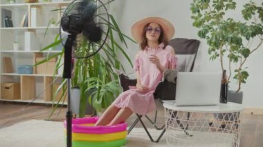Güneş gözlüklü ve plaj şapkalı gülümseyen genç kadın ev bitkilerinin arasında çalışıyor ve tatilin hayalini kuruyor, dizüstü bilgisayarda çalışan kadın koltukta oturuyor ve kola içiyor.