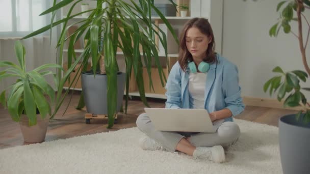 悲しい若い女性が花の間の床のラップトップに座っています 植物は気分と幸福を向上させます 女性は自宅で働きます 女性はノートパソコンで自宅で勉強 女性はノートパソコンでプロジェクトを準備しています — ストック動画