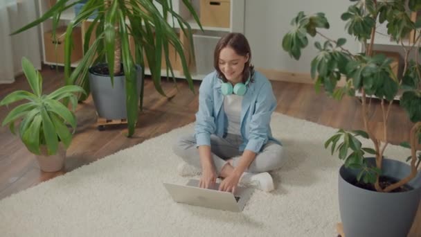 微笑的年轻女子坐在花坛地板上的笔记本电脑前 植物改善心情和幸福 妇女在家工作 妇女在家与手提电脑一起学习 妇女正在准备一个手提电脑项目 — 图库视频影像