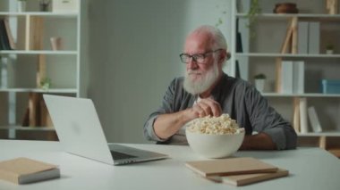 Gülümseyen yaşlı adam dizüsten film izliyor ve patlamış mısır yiyor. Neşeli Yaşlı Adam Komik Bir Film İzlerken Güler. Bir Rahatlama ve Psikolojik Rahatlama Biçimi olarak Filmler.