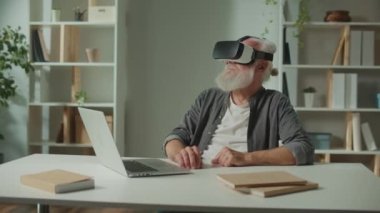 VR gözlüklü, gri sakallı, modern bir yaşlı adam çevredeki her şeyi inceler. VR gözlüklü yaşlı bir adam elinde dizüstü bilgisayarla masada oturuyor. Yaşlılar için sanal gerçeklik eğitimi..