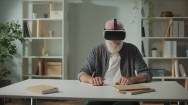 VR gözlüklü, gri sakallı, modern bir yaşlı adam çevredeki her şeyi inceler. VR gözlüklü yaşlı bir adam yaşlılara yardım etmek için Home.VR 'daki masada oturuyor..