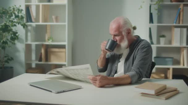 Serious Smart Old Manはテーブルに座り 新聞を読む 新聞を持つ高齢者 自宅でコーヒーを飲む 新聞でニュースと現在のイベントを見る毎日の新聞のコンセプト — ストック動画