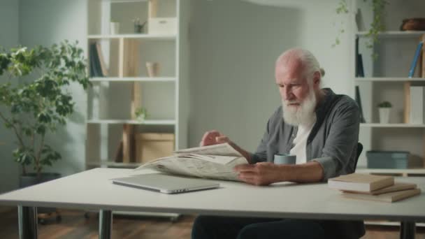 一个严肃 聪明的老人坐在桌旁阅读报纸 一个拿着报纸的老年人 一个人在家喝咖啡 可以看到报纸上的新闻和时事 — 图库视频影像